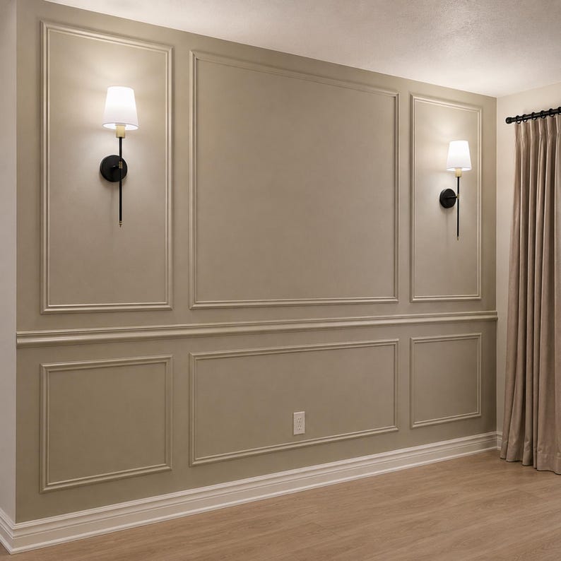 May include: A beige wall featuring decorative rectangular molding panels and two wall-mounted sconces with black metal stems and white fabric shades. The wall is accented by a horizontal chair rail, white baseboards, and light-colored wood flooring.