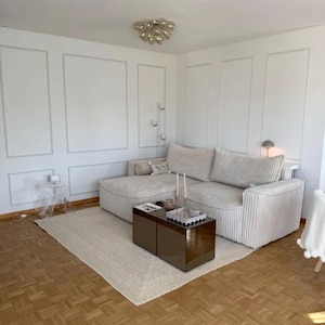 May include: A modern living room with a large, cream-colored sectional sofa, a rectangular coffee table, and a light-colored rug. The walls are white with decorative molding. A clear side table and a decorative light fixture are also visible.