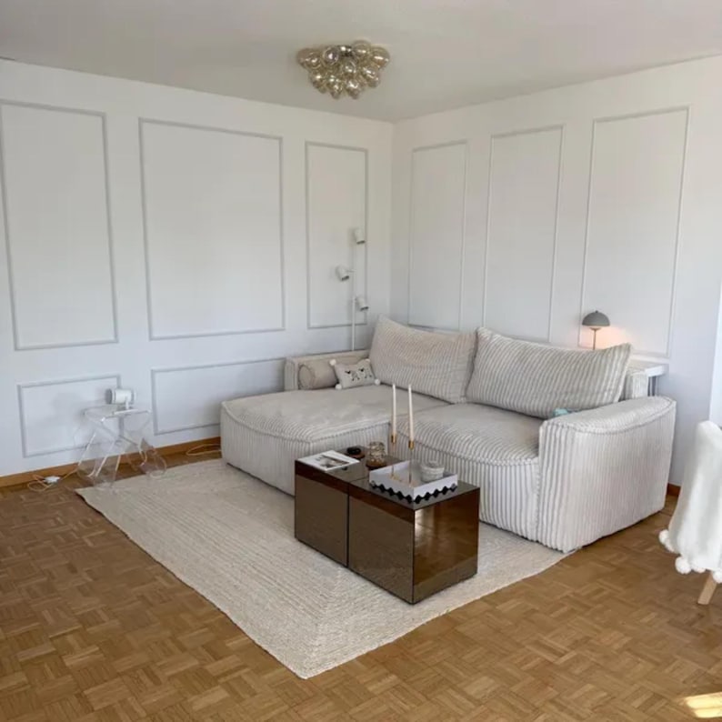 May include: A modern living room with a large, cream-colored sectional sofa, a rectangular coffee table, and a light-colored rug. The walls are white with decorative molding. A clear side table and a decorative light fixture are also visible.