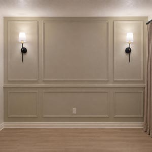 May include: A beige wall featuring decorative rectangular molding panels arranged in a grid pattern. Two wall-mounted light fixtures with black metal arms and white fabric shades are positioned symmetrically on the upper side panels of the wall.