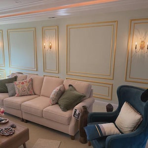 May include: A light beige three-cushion sofa with olive green and floral patterned throw pillows, positioned next to a dark teal velvet wingback chair with a cream pillow, set against a wall with gold-trimmed rectangular molding and wall sconces.