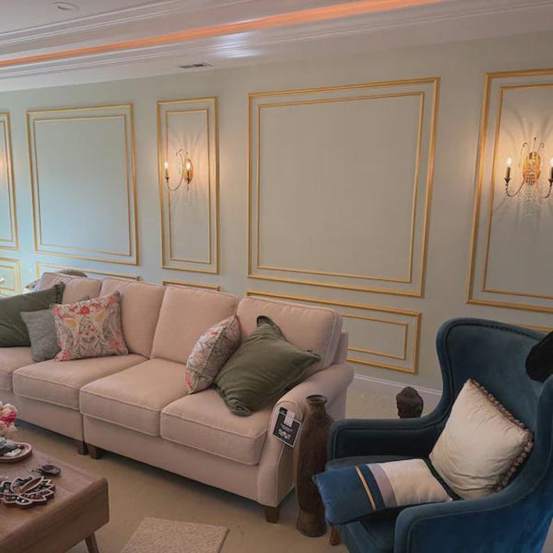 May include: A light beige three-cushion sofa with olive green and floral patterned throw pillows, positioned next to a dark teal velvet wingback chair with a cream pillow, set against a wall with gold-trimmed rectangular molding and wall sconces.