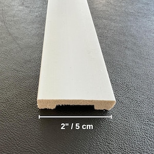May include: A white painted wooden trim piece, 2 inches wide, with a decorative edge. The trim is shown against a dark background, highlighting its clean lines and fresh appearance. Ideal for home improvement projects.