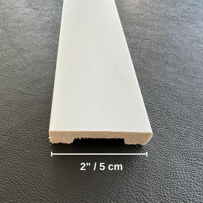 May include: A white painted wooden trim piece, 2 inches wide, with a decorative edge. The trim is shown against a dark background, highlighting its clean lines and fresh appearance. Ideal for home improvement projects.