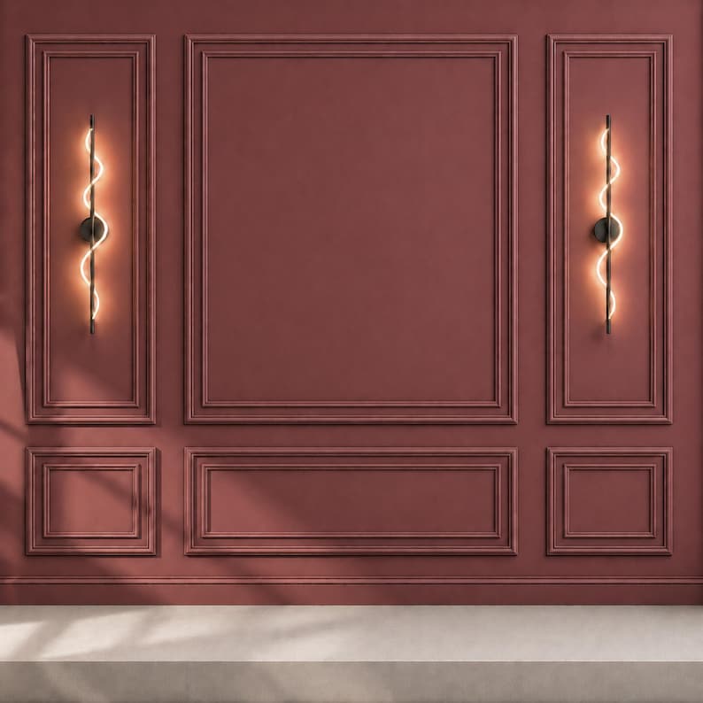 May include: A deep red wall featuring rectangular molding panels and two vertical black wall sconces. Each sconce consists of a straight black rod with a glowing, wavy, illuminated light element running along its length, casting a warm light.