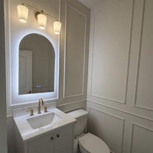 May include: A modern bathroom vanity set with a white sink, gold faucet, and a matching cabinet. Above the sink is a backlit arched mirror and a three-light fixture. The walls are painted a light gray with white trim.