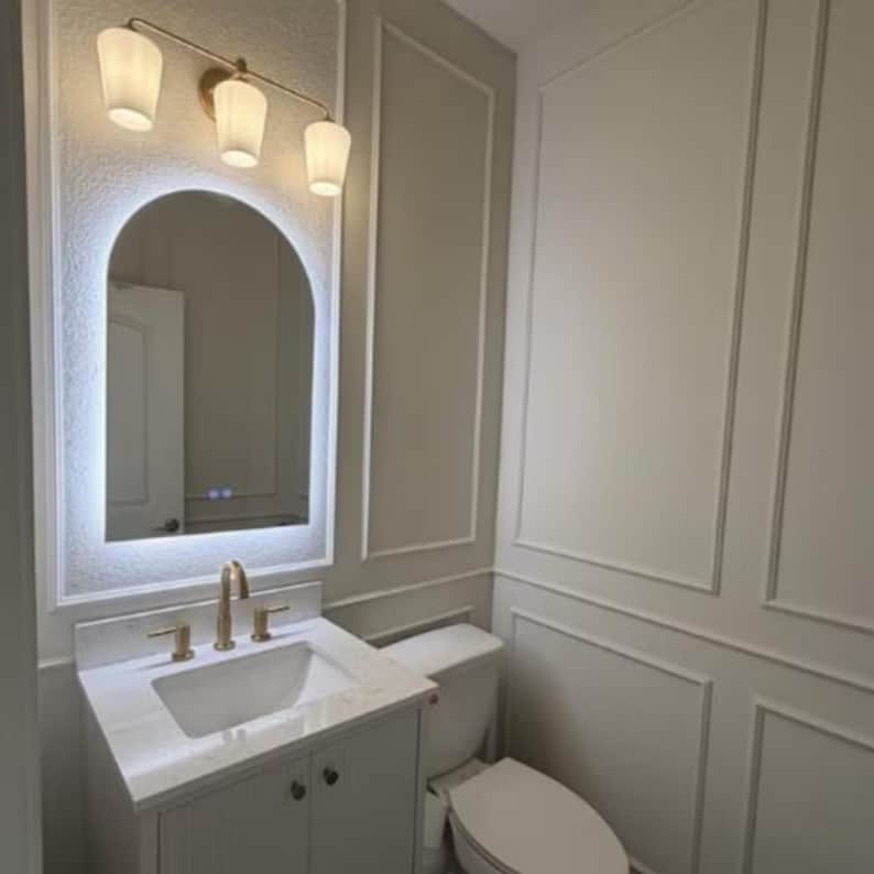 May include: A modern bathroom vanity set with a white sink, gold faucet, and a matching cabinet. Above the sink is a backlit arched mirror and a three-light fixture. The walls are painted a light gray with white trim.