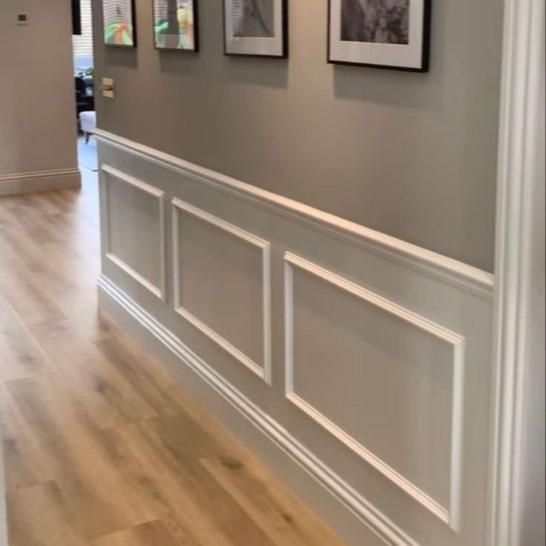 May include: A hallway with white wainscoting and a light gray wall. The wainscoting has a raised panel design with a white trim. There are three framed pictures on the wall above the wainscoting.