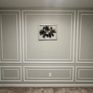 May include: A rectangular wooden window frame wall decoration featuring a central arrangement of dark green pine needles, silver-toned foliage, and a cluster of white and metallic gold spherical ornaments mounted on a light gray wall with molding.