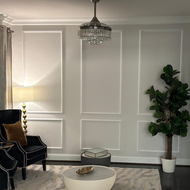 May include: A room interior featuring a ceiling fan with a crystal chandelier light fixture, a black armchair with a gold pillow, a floor lamp with a gold stacked-sphere base, a round coffee table with a gold leaf dish, and a tall potted plant.
