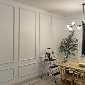May include: A room interior featuring white walls with rectangular molding panels, a wooden dining table with chairs, a black step ladder, a potted artificial olive tree, and a modern black chandelier with multiple spherical light bulbs.