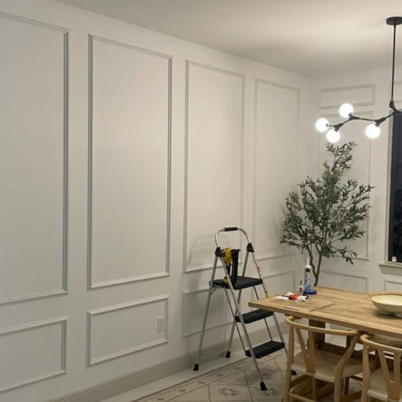 May include: A room interior featuring white walls with rectangular molding panels, a wooden dining table with chairs, a black step ladder, a potted artificial olive tree, and a modern black chandelier with multiple spherical light bulbs.