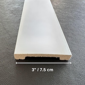 May include: A white painted wooden trim piece, 3 inches wide, with a beveled edge. The text "3" / 7.5 cm" is printed in black below the trim. The trim is likely for home improvement or woodworking projects.