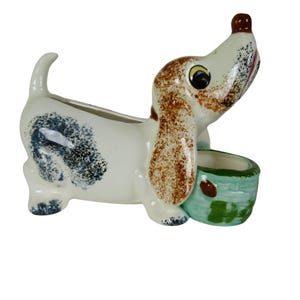 May include: A ceramic dog figurine with a white body, brown and black spots, and a light blue basket. The dog has brown ears, a brown nose, and a smiling expression. The figurine is a decorative item.