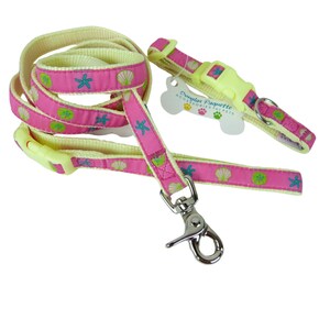 May include: A pink and cream dog leash and collar set. The pink fabric is patterned with seashells, starfish, and green accents. The set includes a leash with a metal clasp and a matching collar with a plastic buckle. A bone-shaped tag reads "Douglas Paquette ACCESSORIES FOR PETS."