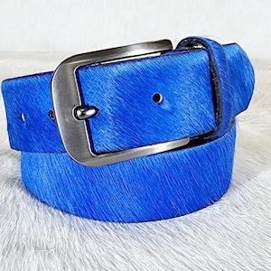 May include: A vibrant blue belt with a silver-toned buckle. The belt is made of a textured material, possibly animal hide, and is displayed on a white, fluffy surface. The buckle is a simple, rectangular design.