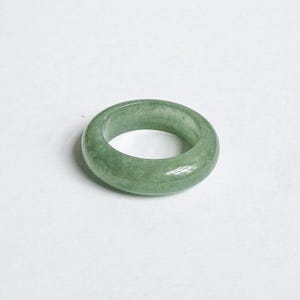 May include: A smooth, round jade ring in a light green hue. The ring is a solid band with a polished surface, showcasing the natural variations in the stone. The ring's design is simple and classic, suitable for everyday wear.