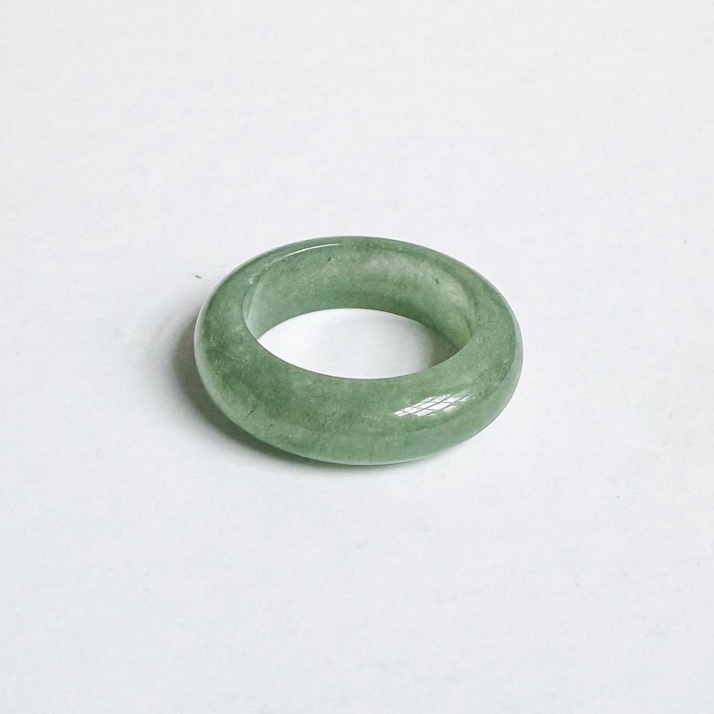 May include: A smooth, round jade ring in a light green hue. The ring is a solid band with a polished surface, showcasing the natural variations in the stone. The ring's design is simple and classic, suitable for everyday wear.