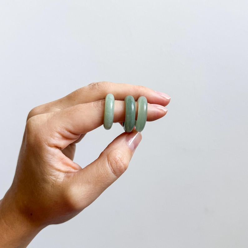 May include: Three jade rings in varying shades of green are displayed. The rings are smooth and polished, with a rounded shape. The rings are held up against a plain white background.