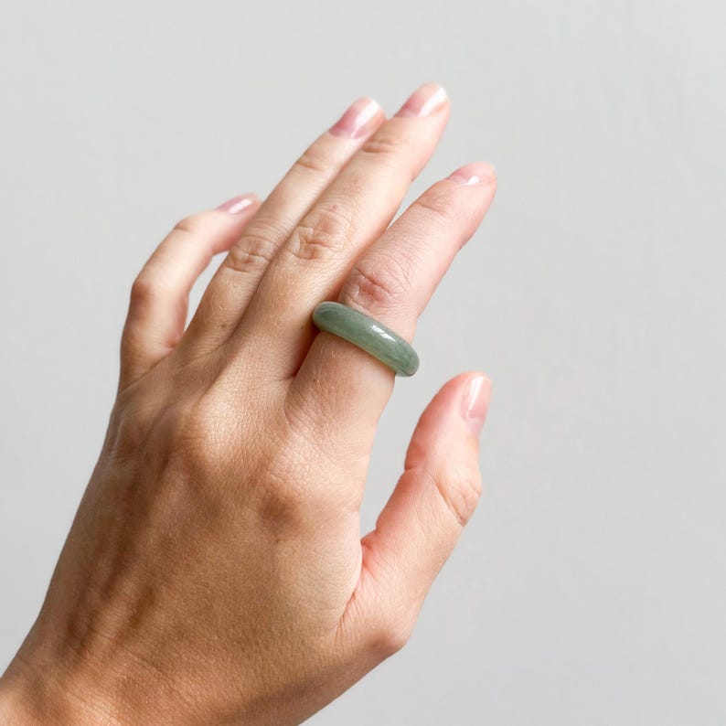 May include: A light green jade ring. The ring is a solid band of polished jade, showcasing a smooth, rounded design. The jade has a subtle, translucent quality, with variations in the green hue. The ring is worn on a finger.