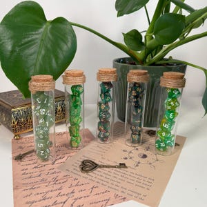 Könnte beinhalten: Fünf Glasfläschchen gefüllt mit grünen und goldenen Würfeln, jeweils mit Korken und Bindfaden verschlossen. Die Würfel werden auf Vintage-Papier mit einem Schlüssel und einer dekorativen Box präsentiert. Eine grüne Pflanze ist ebenfalls sichtbar.