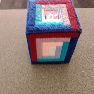 May include: A square tissue box cover made of fabric with a colorful geometric pattern. The cover is made of blue, red, pink, and teal fabric.