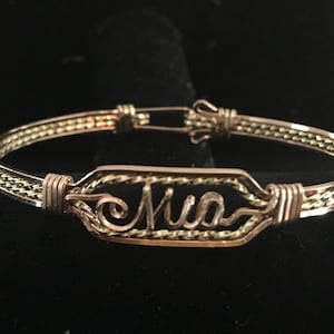 May include: A gold wire bracelet with the name "Mia" inscribed on a rectangular tag. The bracelet is made of two different types of wire, one gold and one silver, twisted together.