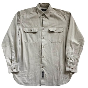 Puede incluir: Camisa de color beige claro de manga larga con dos bolsillos en el pecho y botones. La camisa tiene un cuello clásico y puños abotonados. Una pequeña etiqueta es visible cerca del dobladillo inferior. La camisa es de tela lisa.