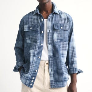 May include: A long-sleeve button-down shirt featuring a blue patchwork design with various plaid and striped patterns. The shirt includes a standard collar, two chest pockets with button flaps, and white buttons down the front placket and cuffs.