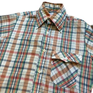 May include: A vintage plaid shirt in brown, red, green, and blue. The shirt has a pointed collar, a chest pocket, and white buttons. The fabric appears to be a lightweight cotton or blend. The shirt is a classic style.
