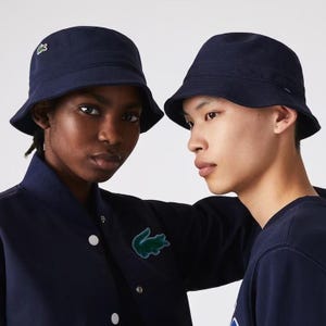 May include: Two navy blue bucket hats with a small green crocodile logo on the side. The hats are worn by two people, one wearing a navy blue bomber jacket with a green crocodile logo and the other wearing a navy blue sweatshirt.