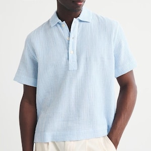 May include: A short-sleeved polo shirt featuring a light blue and white vertical pinstripe pattern. The shirt has a classic collar, a partial button-down placket with three visible white buttons, and a straight hemline with a textured fabric finish.
