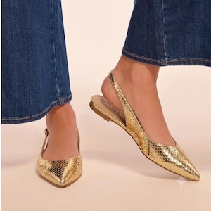 Peut inclure: Une paire de chaussures plates à bride arrière, de couleur dorée métallique, avec un bout pointu et un motif texturé en peau de serpent. Les chaussures présentent une fine bride au talon et la marque Stuart Weitzman est imprimée en doré.