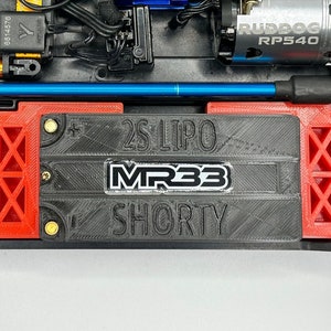 Op de afbeelding: Een zwarte batterijbak met rode zijsteunen. De bak heeft de tekst "2S LIPO MR33 SHORTY" erop gedrukt.