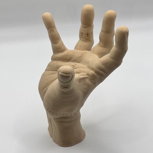 Puede incluir: Una escultura de mano de madera beige con detalles cosidos en los dedos. La mano est&aacute; levantada con los dedos extendidos.