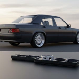 Może przedstawiać: Czarna konsola do wnętrza samochodu z uchwytem na kubek i schowkami. Konsola jest czarna i stoi na czarnej powierzchni. W tle ciemnoszary sedan.