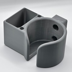 Może przedstawiać: Szary uchwyt ścienny wydrukowany w 3D z uchwytem na kubek i prostokątnym schowkiem. Uchwyt na kubek ma zaokrąglony otwór i dwa otwory z boku. Schowek ma dwa otwory na górze.