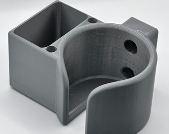 Puede incluir: Un soporte de pared gris impreso en 3D con un portavasos y un compartimento de almacenamiento rectangular. El portavasos tiene una abertura redondeada y dos agujeros en el lateral. El compartimento de almacenamiento tiene dos agujeros en la parte superior.