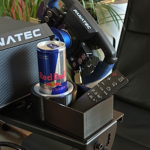 Könnte beinhalten: Schwarzer Rennsimulator-Ständer mit einem schwarzen Lenkrad, einer Dose Red Bull Energy Drink und einer schwarzen Fernbedienung. Das Lenkrad trägt das Fanatec-Logo. Der Ständer ist in Deutschland hergestellt.