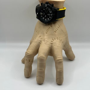 Puede incluir: Un reloj de pulsera negro con una correa amarilla se muestra en un soporte en forma de mano beige. La esfera del reloj tiene agujas negras y marcas blancas.