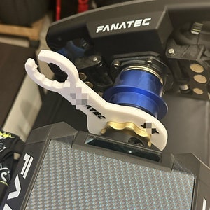 Könnte beinhalten: Ein weißes Plastikwerkzeug mit dem Aufdruck "Fanatec". Das Werkzeug dient zum Einstellen des Lenkrads eines Rennsimulators. Das Werkzeug liegt auf einer schwarzen Basis mit einem Karbonfaser-Muster.