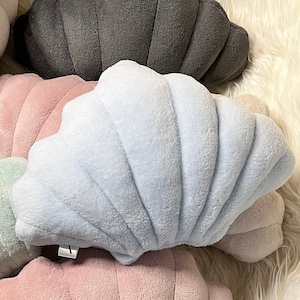 Könnte beinhalten: Eine Sammlung von plüschigen, croissantförmigen Kissen in verschiedenen Pastellfarben, darunter Hellblau, Rosa, Grau und Mintgrün. Die Kissen haben eine weiche, strukturierte Oberfläche und sind auf einer weißen, flauschigen Oberfläche angeordnet.