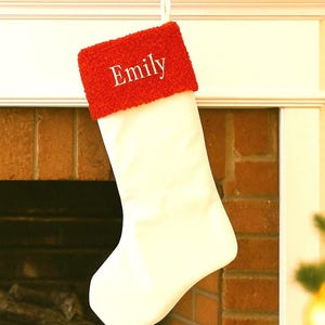 Könnte beinhalten: Ein weißer Weihnachtsstrumpf mit roter Manschette, personalisiert mit dem Namen "Emily" in weißer Schrift. Der Strumpf hängt an einem goldenen Haken über einem Ziegelkamin, der mit Grün geschmückt ist.