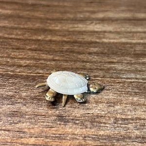 Puede incluir: Pequeña figura de tortuga con caparazón blanco y cuerpo color bronce. La tortuga descansa sobre una superficie de madera, mostrando su intrincado diseño y textura. La figura es un objeto decorativo.