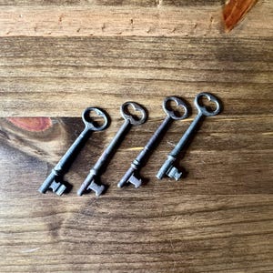 May include: Four antique skeleton keys with ornate tops and detailed key bits are arranged diagonally on a wooden surface. The keys have a dark, aged metal finish, suggesting they are vintage or vintage-inspired decorative items.