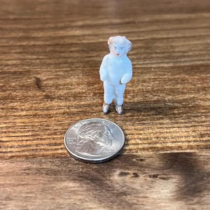 May include: A small, antique porcelain doll figurine, painted in white with silver shoes. The doll has a detailed face with painted features. A U.S. quarter coin is next to the doll for size comparison. The doll is standing on a wooden surface.