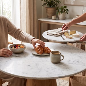 May include: A round white marble dining table with a light wood base. On the table sits a ceramic bowl filled with fresh fruit, a small plate with two golden-brown croissants, and a speckled ceramic mug containing a light brown beverage.