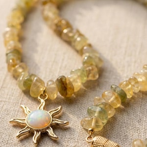 May include: A gold-colored necklace with a sun pendant and a strand of yellow and green gemstone beads. The sun pendant has a white opal center. The necklace is displayed on a neutral linen surface.