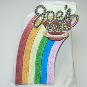 Op de afbeelding: Geborduurde patch met een regenboogontwerp en de tekst "Joe's Cafe". De patch heeft een koffiekopje en is in tinten rood, oranje, geel, groen, blauw en paars. Geschikt voor kleding of accessoires.