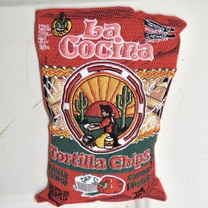 May include: An embroidered patch depicting a bag of tortilla chips with the text "La Cocina Tortilla Chips". The bag features a cartoon image of a person making tortillas and the text "Fresh Flavour" and "Savour Fresh".